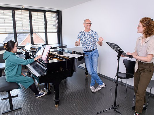 Unterrichtssituation mit einer Lehrkraft und zwei Studierenden mit Flügel und Notenpult